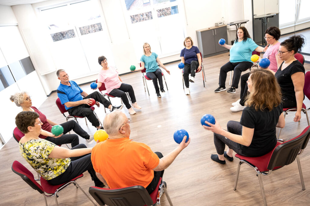 Eine gemischte Gruppe sitzt auf Stühlen im Kreis mit bunten Bällen in einer Hand – Reha-Sport-Kurs Hocker