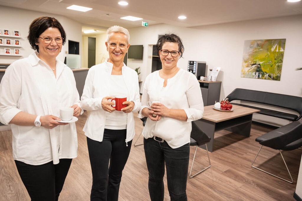 Drei Mitarbeiterinnen von Sport & Therapiezentrum Schumann im Empfangsbereich mit einer Tasse in der Hand